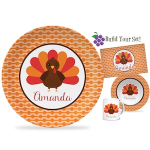 May include: A Thanksgiving themed children's dinnerware set with a turkey design. The set includes a plate, bowl, mug, and placemat. The items are orange and white with a turkey design and the name "Amanda" printed on each piece.