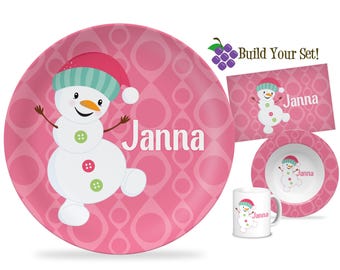 Könnte beinhalten: Ein rosa Schneemann-Geschirr-Set mit einem Teller, einer Schüssel, einem Platzset und einer Tasse. Jedes Teil hat einen rosa Hintergrund mit einem sich wiederholenden Muster und einem lächelnden Schneemann mit Hut. Der Name "Janna" ist aufgedruckt.