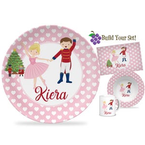 May include: A pink dinnerware set featuring a ballerina and a soldier design with the name "Kiera". The set includes a plate, bowl, placemat, and mug. The plate and placemat have a white heart pattern.