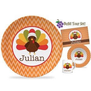 May include: Thanksgiving-themed dinnerware set featuring a cartoon turkey wearing a Santa hat. The set includes a large plate, a smaller bowl, a mug, and a placemat, all with the name "Julian". The background has an orange chevron pattern.