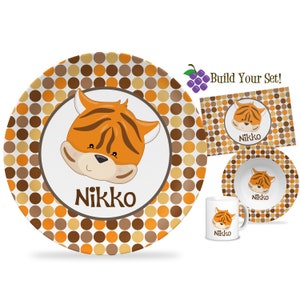 May include: Personalized children's dinnerware set with a tiger design. The set includes a plate, bowl, and mug. The plate and bowl have a brown and orange polka dot pattern. The mug has a brown and orange polka dot pattern with a tiger face on the front. The set is personalized with the name "Nikko".