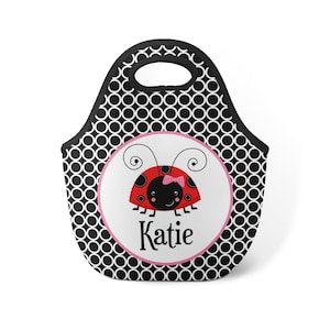 May include: Black and white polka dot lunch bag with a pink circle featuring a red ladybug with black spots and a pink bow. The name "Katie" is written in black letters below the ladybug.
