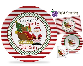 Könnte beinhalten: Ein weihnachtliches Geschirr-Set mit einem großen Teller mit rot-weißen Streifen, einem kleineren Teller, einer Tasse und einem rechteckigen Tablett. Jedes Teil hat ein zentrales Design von Santa Claus und einem Schlitten mit einem Teddybär und Geschenken. Text: "Cookies for Santa" und "Love, Jordan."