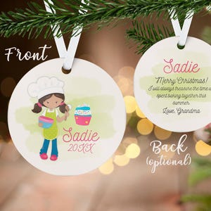 May include: A round, white Christmas ornament with a cartoon illustration of a girl baker holding a bowl and spoon. The ornament has the name "Sadie" and the year "20XX" on the front. The back says "Merry Christmas!" and a message from "Love, Grandma."