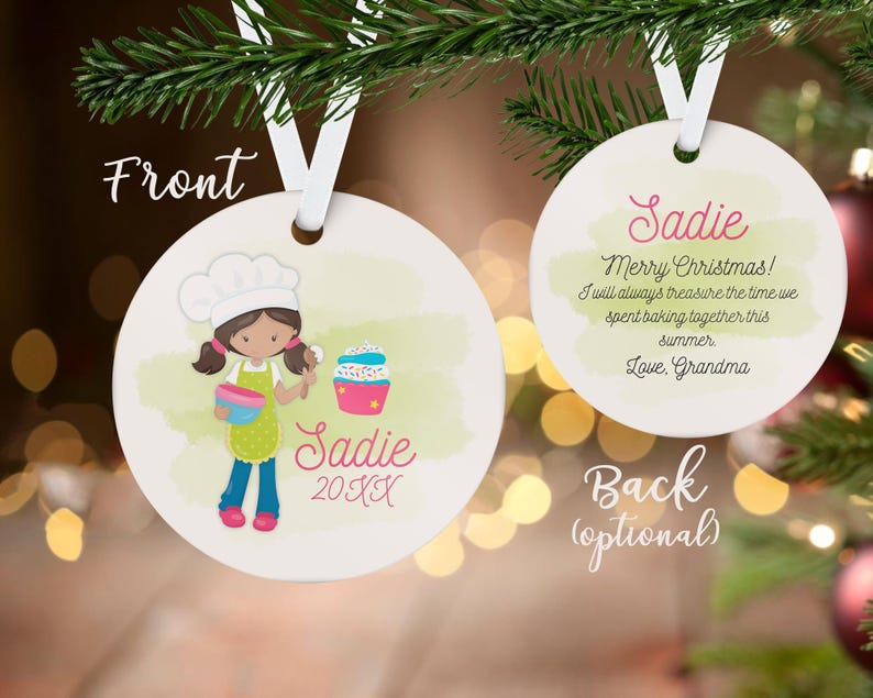 May include: A round, white Christmas ornament with a cartoon illustration of a girl baker holding a bowl and spoon. The ornament has the name "Sadie" and the year "20XX" on the front. The back says "Merry Christmas!" and a message from "Love, Grandma."