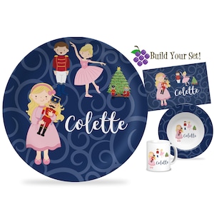 May include: A children's dinnerware set featuring a navy blue plate with a ballerina, nutcracker, and Christmas tree design. Includes a matching bowl, mug, and placemat. The name "Colette" is written on each piece.