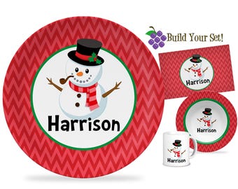 Könnte beinhalten: Ein Set von Schneemann-Geschirr, einschließlich Teller, Schüssel, Tischset und Becher. Jeder Artikel zeigt ein Schneemanndesign mit schwarzem Zylinder, rotem Schal und dem Namen "Harrison". Der Hintergrund hat ein rotes Zickzackmuster.