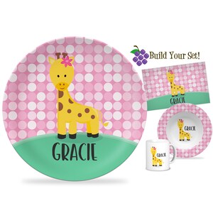 May include: A children's dinnerware set featuring a yellow giraffe with brown spots and a pink flower. The set includes a plate, bowl, placemat, and mug, all with the name "Gracie". The background has a pink and white polka dot pattern.