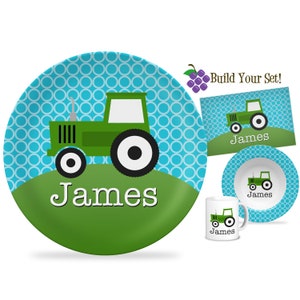May include: A children's dinnerware set featuring a green tractor design. The set includes a plate, bowl, mug, and placemat, all with a blue and white patterned background. The name "James" is printed on each item.