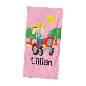 May include: Pink beach towel with a white heart pattern. A cartoon girl is driving a red tractor with a basket of apples. The text "Lillian" is printed on the towel.