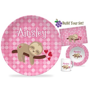 May include: A pink polka-dot plate, placemat, bowl, and mug set featuring a cartoon sloth on a branch with red hearts. The name "Ainsley" is written in white script. The set is designed for children.