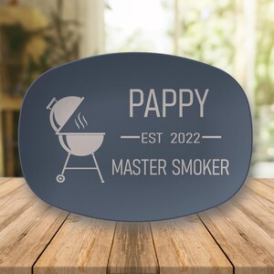 May include: A navy blue oval platter with white text and a grill graphic. The text reads "PAPPY EST 2022 MASTER SMOKER". The platter is on a wooden surface.
