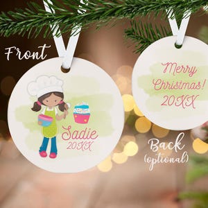 May include: A white, round Christmas ornament with a cartoon illustration of a girl chef holding a bowl and spoon, with a cupcake. The ornament has the name "Sadie" and the year "20XX". The back says "Merry Christmas! 20XX".