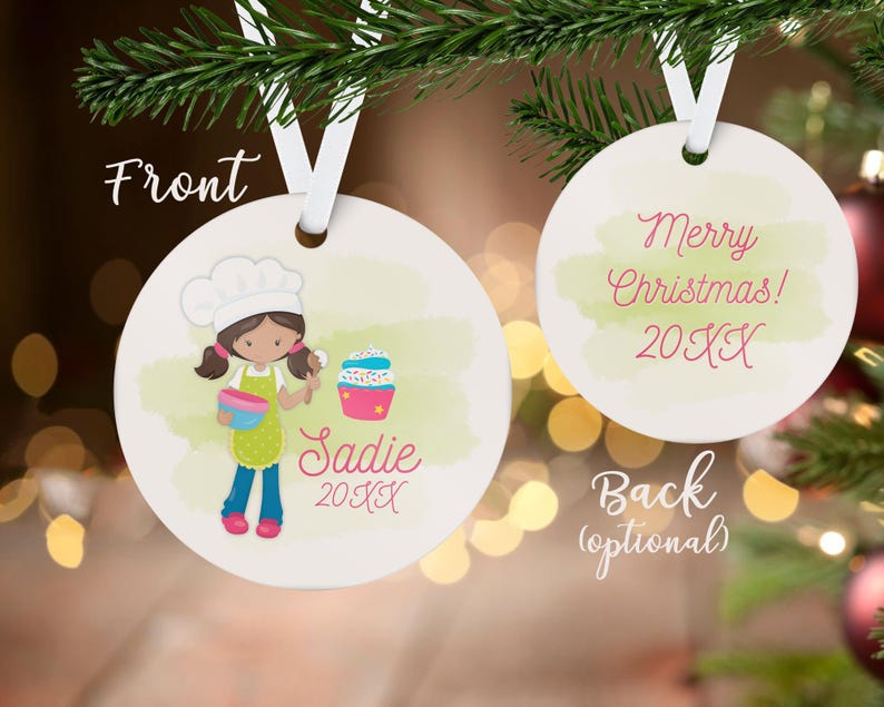May include: A white, round Christmas ornament with a cartoon illustration of a girl chef holding a bowl and spoon, with a cupcake. The ornament has the name "Sadie" and the year "20XX". The back says "Merry Christmas! 20XX".