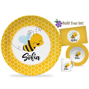 May include: A personalized children's dinnerware set featuring a cute cartoon bee with a honeycomb pattern. The set includes a plate, bowl, and mug, all with the name "Sofia" printed on them. Build Your Set!