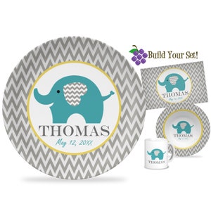 May include: A personalized baby dish set with a gray and white chevron pattern. The set includes a large plate, a small bowl, a mug, and a placemat. Each piece features a blue elephant with a gray chevron pattern on its ear. The name "THOMAS" and the date "May 12, 20XX" are printed on each piece.