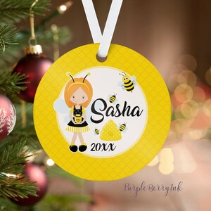 May include: A round, yellow Christmas ornament with a bee-themed design. The ornament features a cartoon girl dressed as a bee, the name "Sasha", and the year "20XX". The ornament is hanging from a white ribbon.