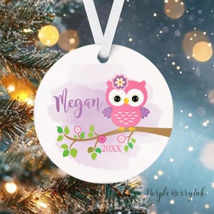 May include: A white, round Christmas ornament with a pink owl perched on a branch. The ornament features the name "Megan" in purple script, along with the year "20XX". A white ribbon is attached for hanging.