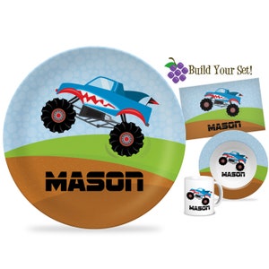 May include: A blue monster truck with red accents and a toothy grin jumps over a green hill. The truck is on a blue and brown plate with the name "MASON" printed on it.  The image also shows a matching bowl, mug, and rectangular plate.