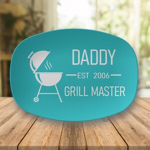 May include: A teal oval platter with white text and a grill graphic. The text reads "DADDY - EST 2006 - GRILL MASTER". The graphic is of a grill with the lid open and smoke rising. The platter is on a wooden surface.