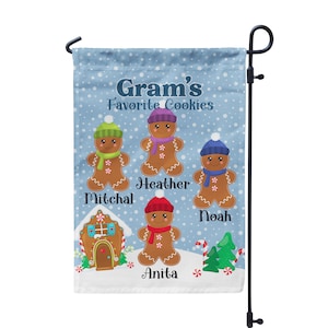 May include: A blue Christmas garden flag with the text "Gram's Favorite Cookies" and four gingerbread people with names: Mitchel, Heather, Noah, and Anita. The gingerbread people are wearing winter hats and scarves. There is a gingerbread house in the background.