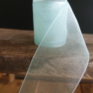May include: A roll of light blue sheer ribbon with a delicate, open weave. The ribbon is partially unrolled, revealing its texture and the cylindrical shape of the roll. The ribbon sits on a wooden surface against a dark background.