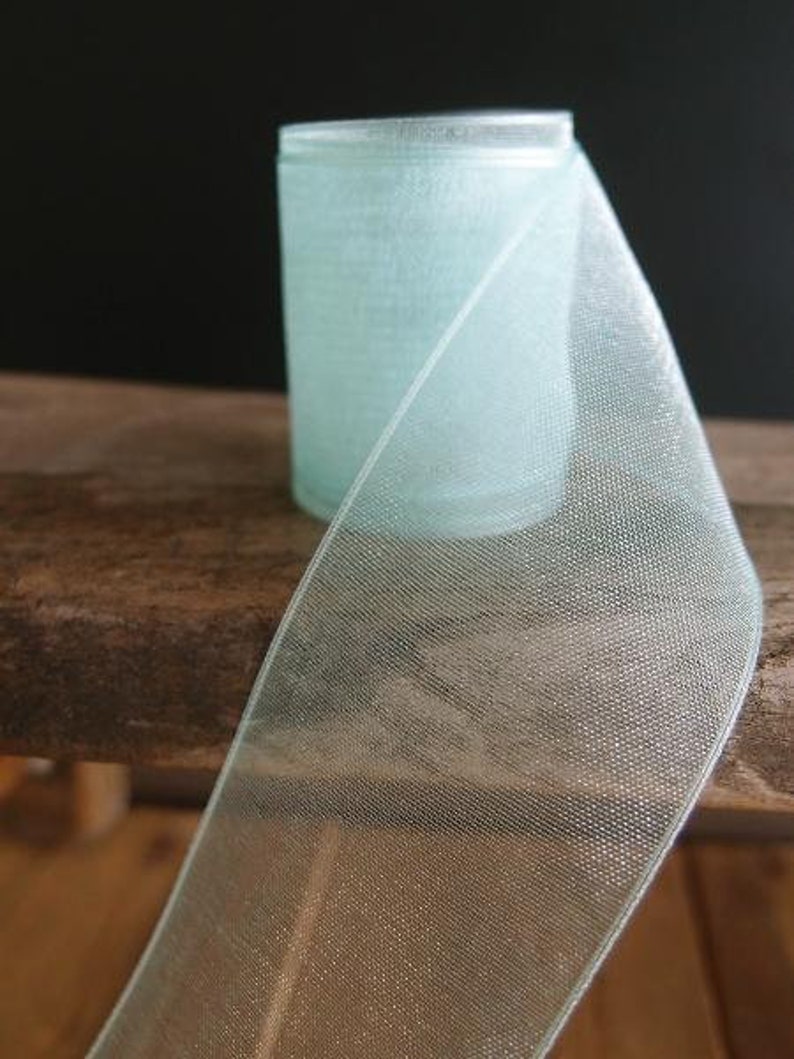 May include: A roll of light blue sheer ribbon with a delicate, open weave. The ribbon is partially unrolled, revealing its texture and the cylindrical shape of the roll. The ribbon sits on a wooden surface against a dark background.