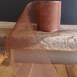 May include: A roll of copper-colored ribbon with a sheer, mesh-like texture. The ribbon is partially unrolled, revealing its width and the tightly wound roll. The ribbon sits on a weathered wooden surface, with a dark background.