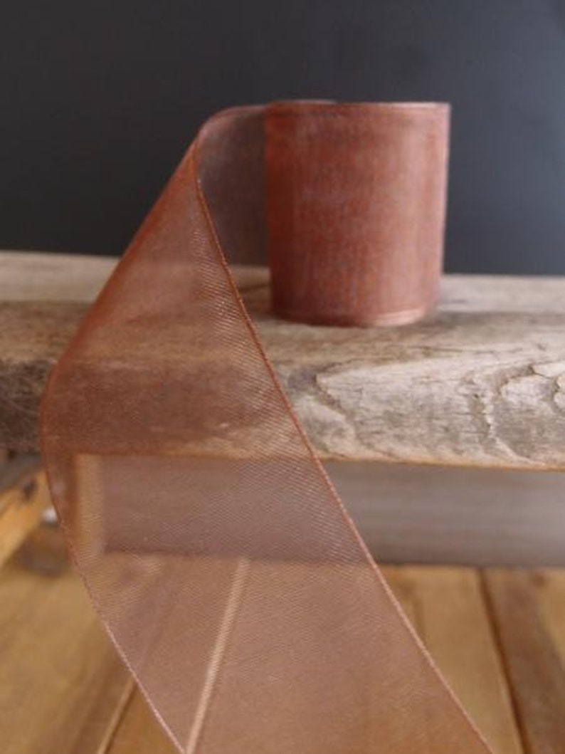 May include: A roll of copper-colored ribbon with a sheer, mesh-like texture. The ribbon is partially unrolled, revealing its width and the tightly wound roll. The ribbon sits on a weathered wooden surface, with a dark background.