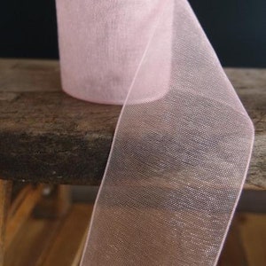 May include: A roll of sheer, light pink ribbon. The ribbon is made of a delicate, open-weave fabric, and a length of the ribbon is unrolled, showing its width. The ribbon is sitting on a wooden surface.