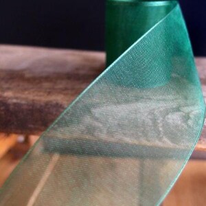 May include: Close-up of a roll of emerald green sheer ribbon. The ribbon has a delicate, open weave texture, and the spool is visible in the background. The ribbon is likely used for crafts, gift wrapping, or decorative purposes.