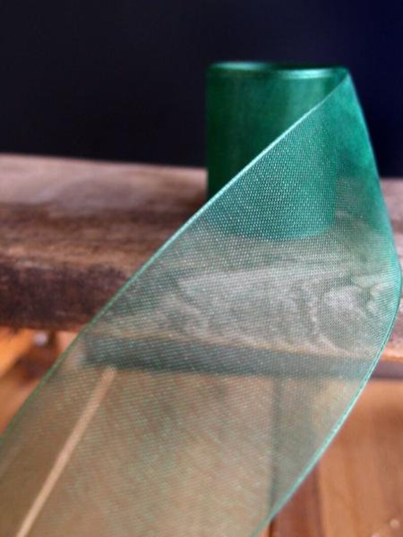 May include: Close-up of a roll of emerald green sheer ribbon. The ribbon has a delicate, open weave texture, and the spool is visible in the background. The ribbon is likely used for crafts, gift wrapping, or decorative purposes.