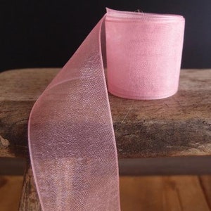 May include: A roll of sheer, light pink ribbon sits on a wooden surface. The ribbon is partially unrolled, revealing its delicate, mesh-like texture. The background is a dark, solid color, highlighting the ribbon's soft hue.