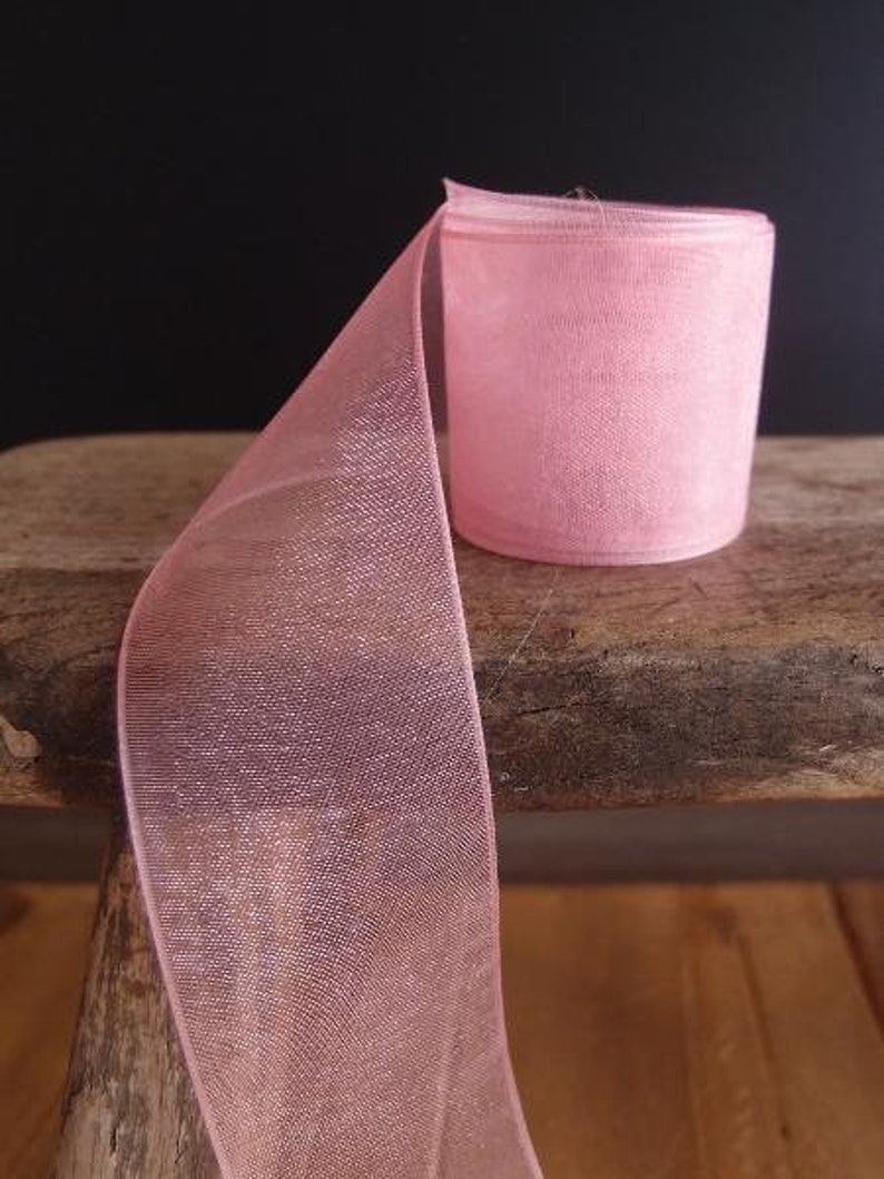 May include: A roll of sheer, light pink ribbon sits on a wooden surface. The ribbon is partially unrolled, revealing its delicate, mesh-like texture. The background is a dark, solid color, highlighting the ribbon's soft hue.