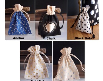 May include: Five drawstring bags made of natural fabric with different patterns. The bags are in a row, from left to right: anchor, chalk, black dots, gold dots, and silver dots.