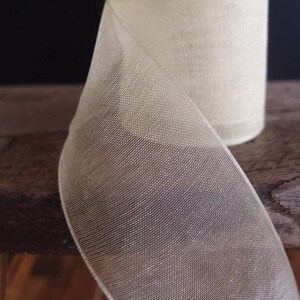 May include: A roll of sheer, ivory-colored ribbon. The ribbon has a delicate, open weave, and a subtle sheen. The spool is partially unrolled, revealing the texture and depth of the material. The ribbon is suitable for crafts, gift wrapping, or decorative purposes.