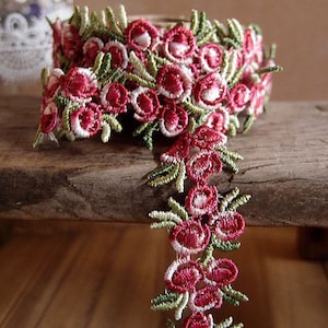 May include: A close-up of a pink and green floral embroidered trim. The trim is made of delicate lace and features small, detailed flowers.