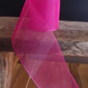 May include: A roll of vibrant fuchsia ribbon. The sheer fabric is folded over, revealing the texture and color of the material. The ribbon is on a wooden surface, with a dark background.