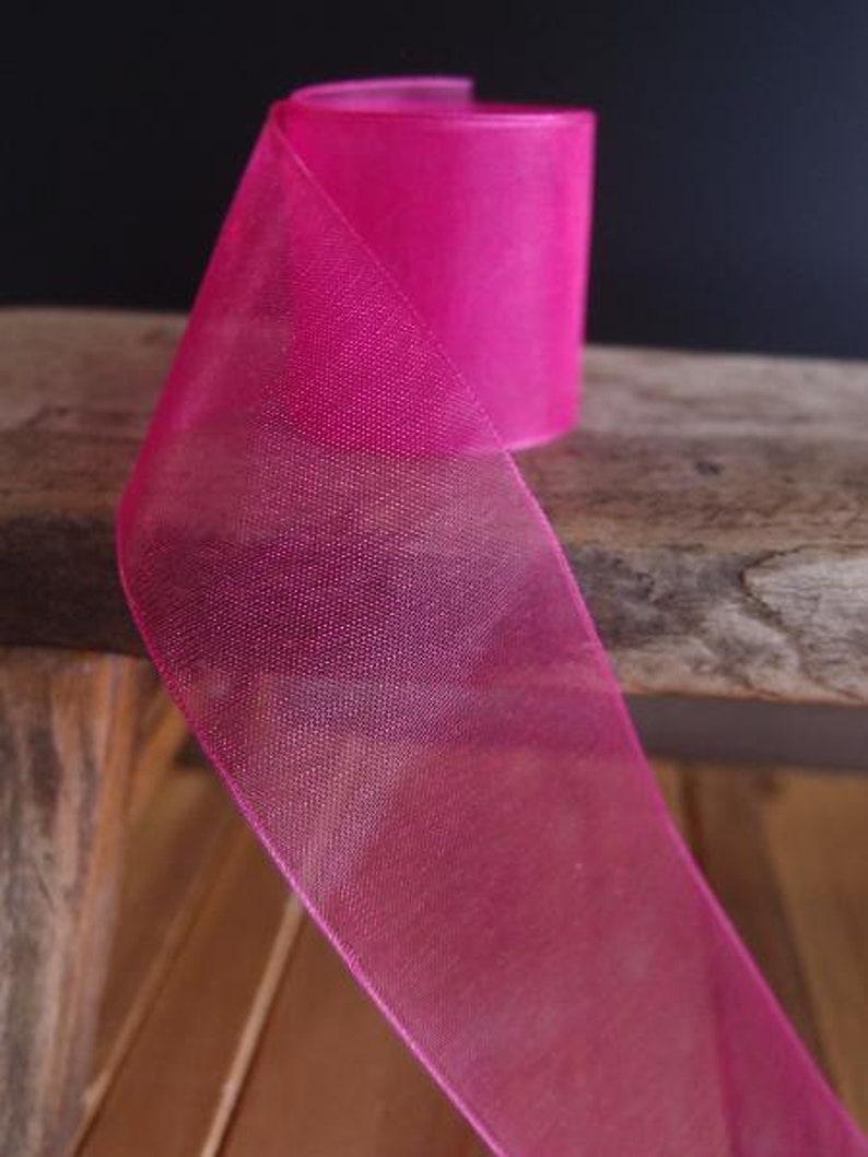 May include: A roll of vibrant fuchsia ribbon. The sheer fabric is folded over, revealing the texture and color of the material. The ribbon is on a wooden surface, with a dark background.
