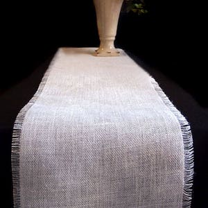 May include: White burlap table runner with frayed edges. The runner is laying flat on a black surface.
