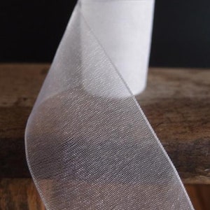 May include: A roll of white, sheer ribbon with a delicate, woven texture. The ribbon is partially unrolled, revealing its translucent quality. The background is a dark, solid color, and the ribbon sits on a wooden surface.