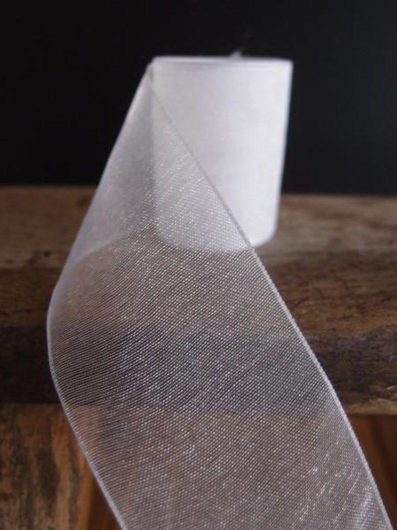 May include: A roll of white, sheer ribbon with a delicate, woven texture. The ribbon is partially unrolled, revealing its translucent quality. The background is a dark, solid color, and the ribbon sits on a wooden surface.
