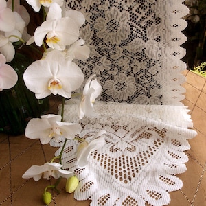 May include: White lace table runner with a delicate floral pattern. The runner is made of a soft, lightweight fabric and has a scalloped edge. The runner is displayed on a brown surface with white orchids.