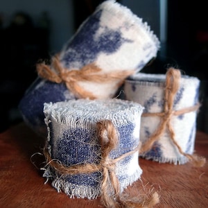 May include: Three rolls of fabric, each tied with twine, are stacked on a wooden surface. The fabric is a blue and white pattern with a frayed edge.
