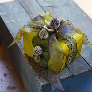 May include: A yellow gift box wrapped with a sheer, olive green and purple ribbon. The box is decorated with several spiky, gray-green floral accents and a blue beaded accent.