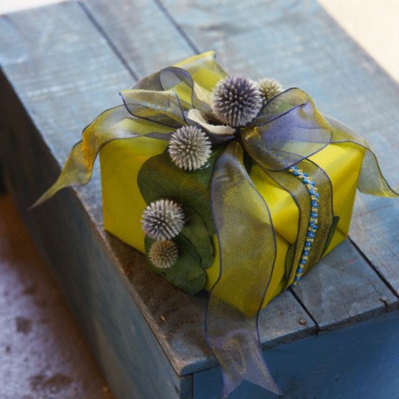 May include: A yellow gift box wrapped with a sheer, olive green and purple ribbon. The box is decorated with several spiky, gray-green floral accents and a blue beaded accent.
