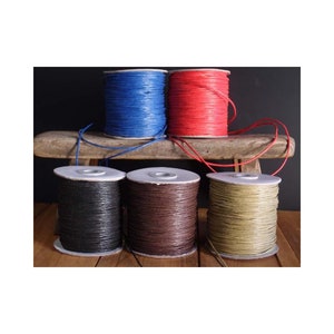 May include: Five spools of waxed cotton cord in different colors: black, brown, beige, red, and blue. The spools are arranged on a wooden surface.