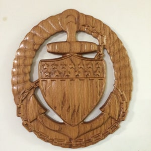 May include: A wooden wall plaque featuring a carved anchor, shield, and rope design. The shield has a carved American flag design with stars and stripes. The anchor is surrounded by a rope design.