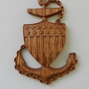 May include: A wooden anchor wall decor with a shield design. The anchor is carved from light brown wood and features a chain, stars, and vertical lines. The design is detailed and has a nautical theme.