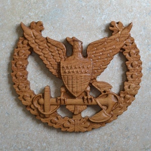 May include: A wooden carving of an eagle with outstretched wings holding an anchor and a crossed axe. The eagle is surrounded by a wreath of oak leaves.
